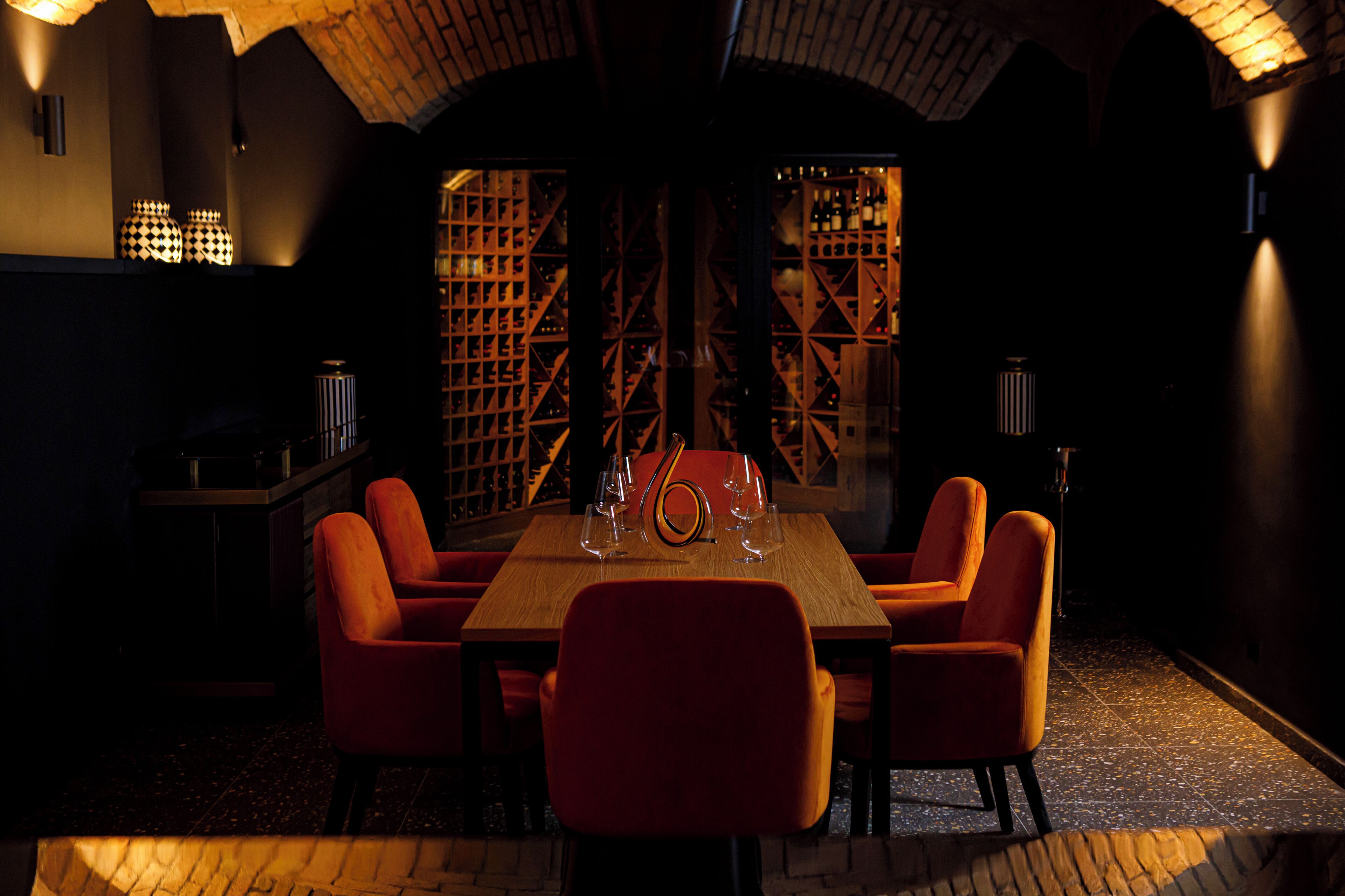 A dimly lit room with a wooden table and orange chairs, a wine rack in the background, and a patterned wall.