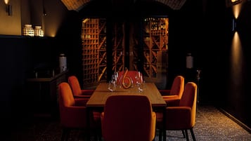 A dimly lit room with a wooden table and orange chairs, a wine rack in the background, and a patterned wall.
