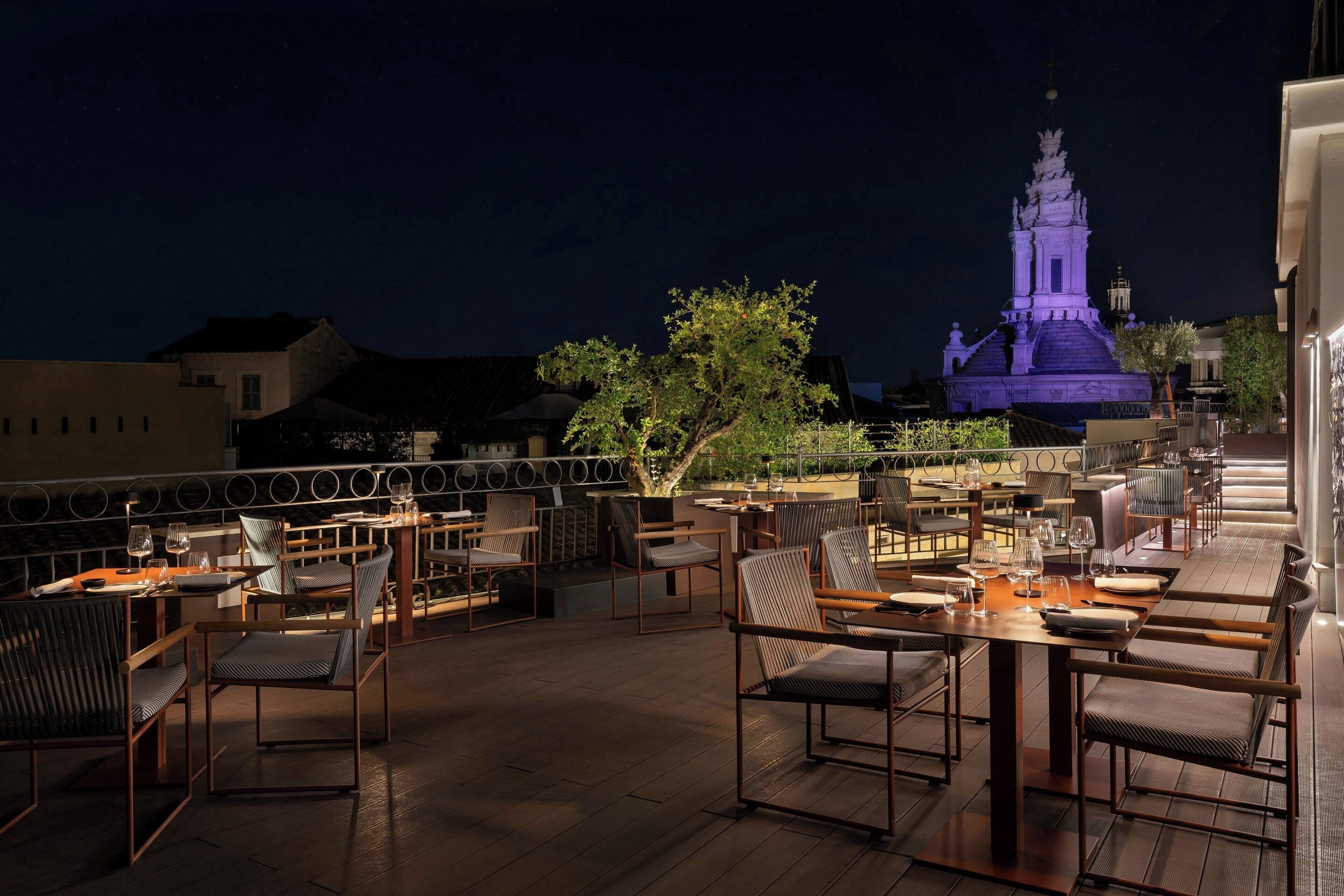 An outdoor dining area with tables and chairs, a tree, and a building with illuminated spires in the background.