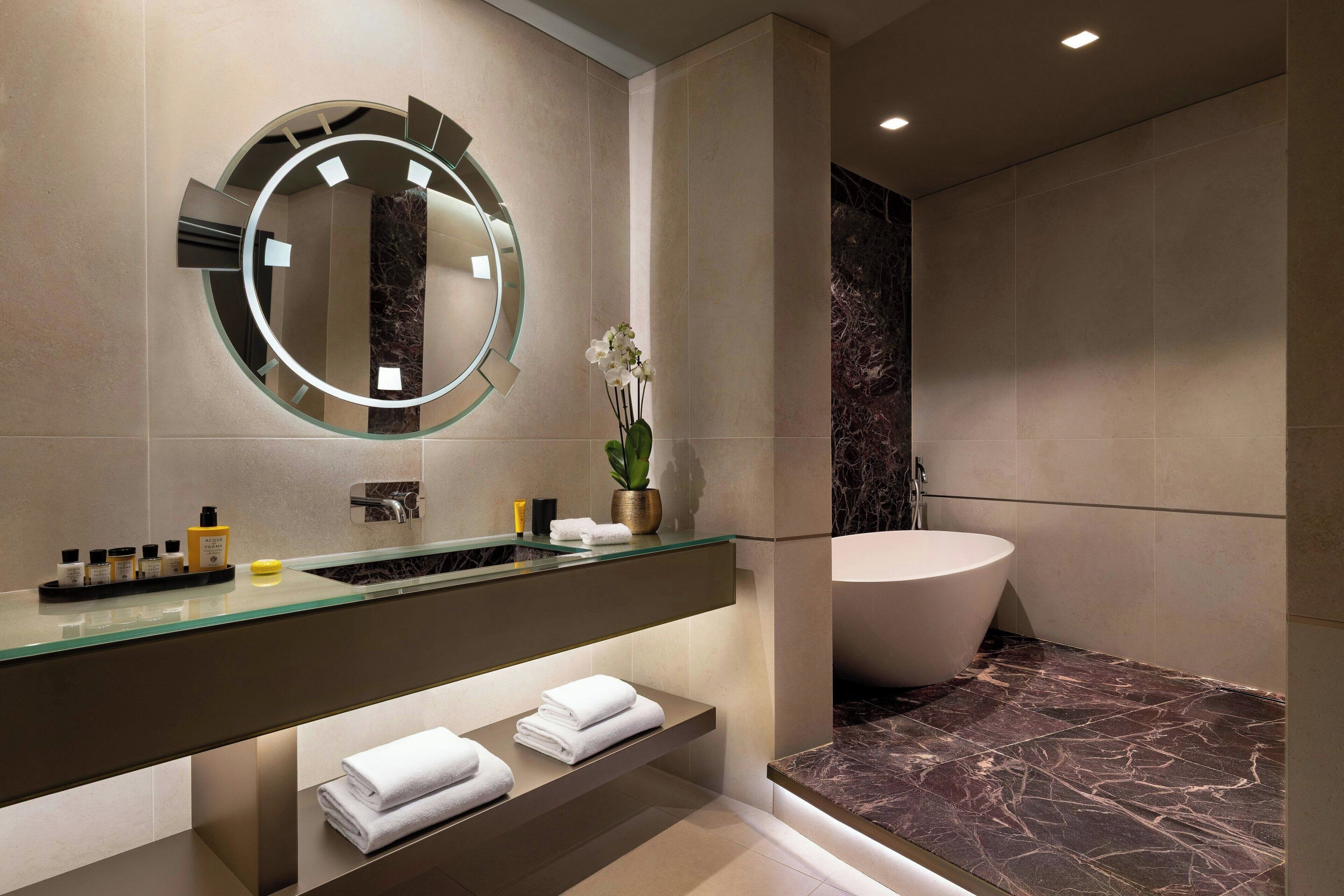 A modern bathroom with a glass vanity, a large round mirror, a bathtub, and a marble floor.