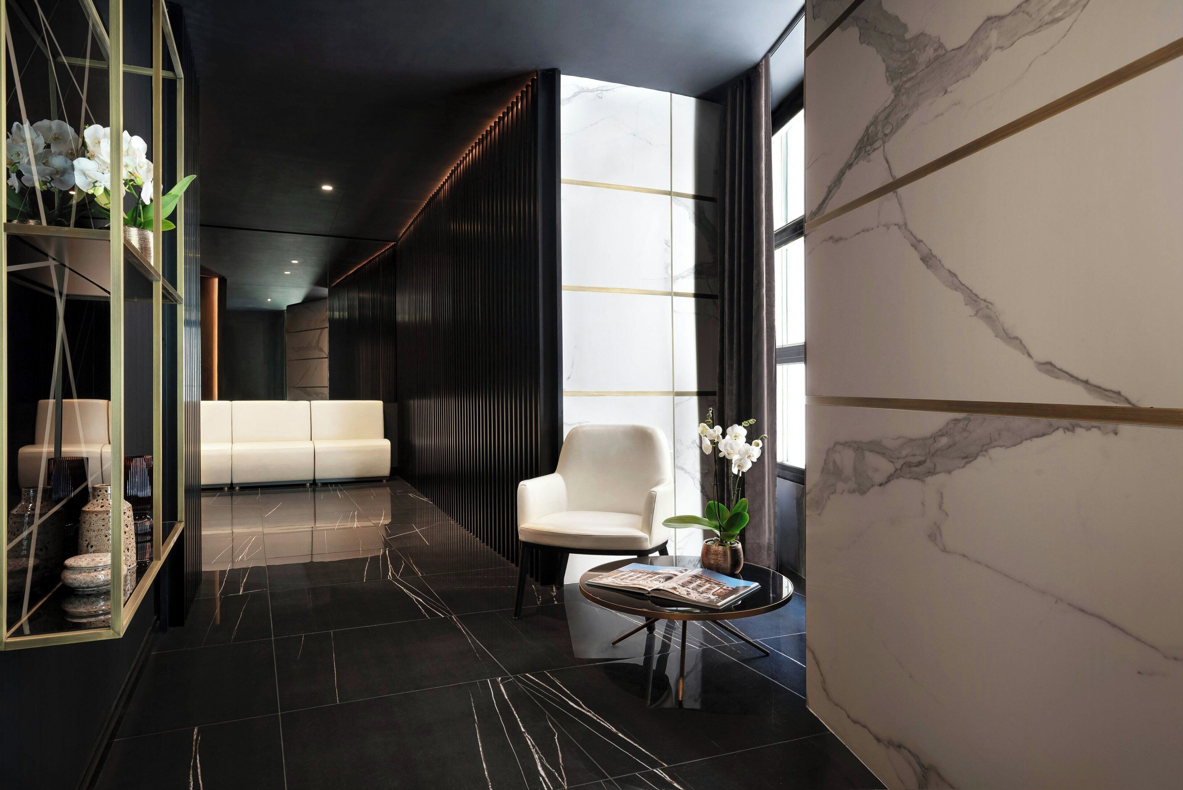 A modern lobby with a marble wall, a white sofa, a round table with a magazine, and a vase of white flowers.