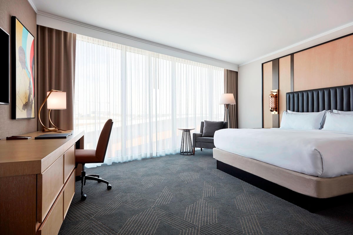 Une chambre d’hôtel avec un lit, un bureau, une chaise, une petite table et une grande fenêtre avec des rideaux.