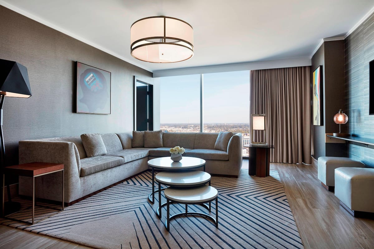 Une chambre d’hôtel moderne, dotée d’un canapé d’angle, d’une petite table basse, d’un lampadaire et d’une grande fenêtre donnant sur la ville.