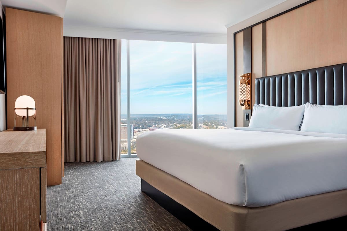 Une chambre d’hôtel avec un grand lit, un bureau, une lampe et une vue sur la ville grâce à de grandes fenêtres.