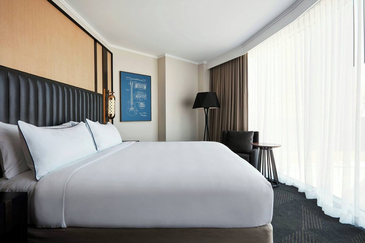 Une chambre d’hôtel avec un grand lit, un bureau, une chaise, une lampe et une fenêtre avec des rideaux.