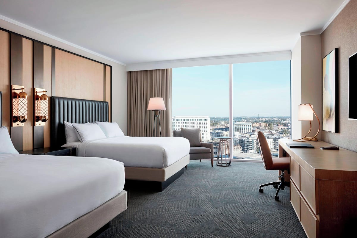 Une chambre d’hôtel avec deux lits, un bureau, une chaise, une lampe et une grande fenêtre donnant sur la ville.