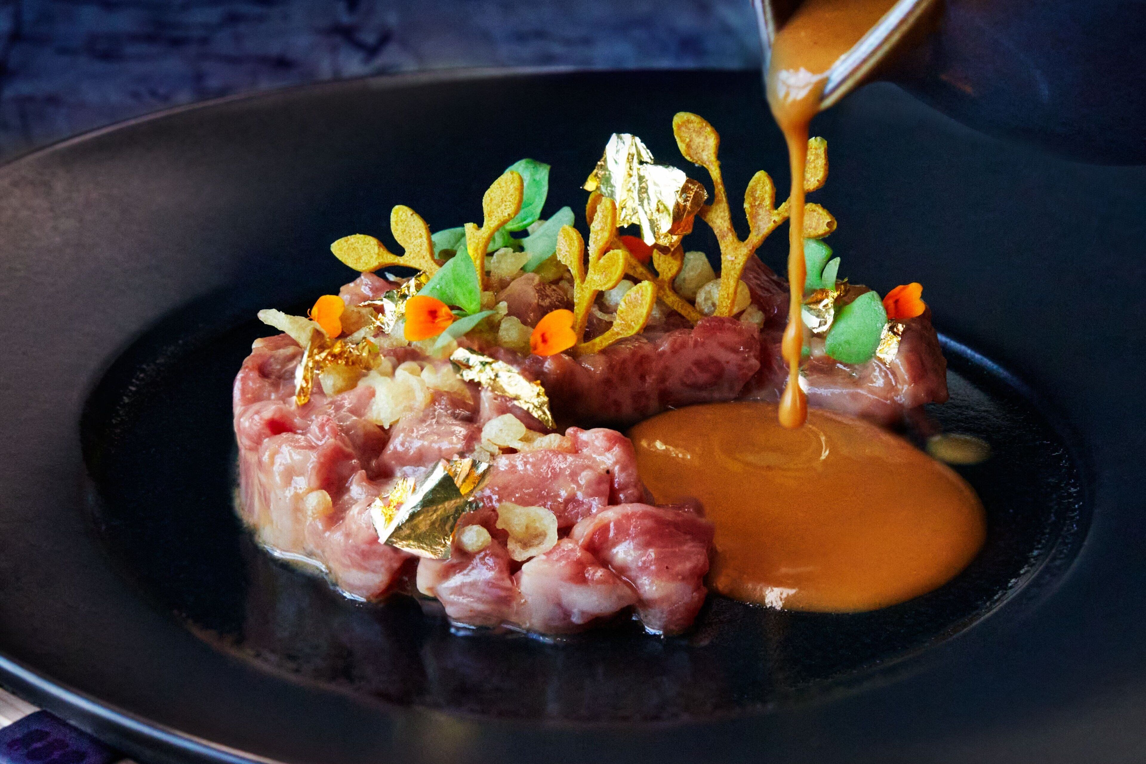 Un plat composé de viande, de légumes et de quelques paillettes d’or, le tout présenté sur une assiette noire.