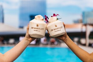 Two hands holding white coconut-shaped containers with "JW MARRIOTT NASHVILLE" text, with a blurred pool and buildings in the background. - JW Marriott Nashville (Nashville)