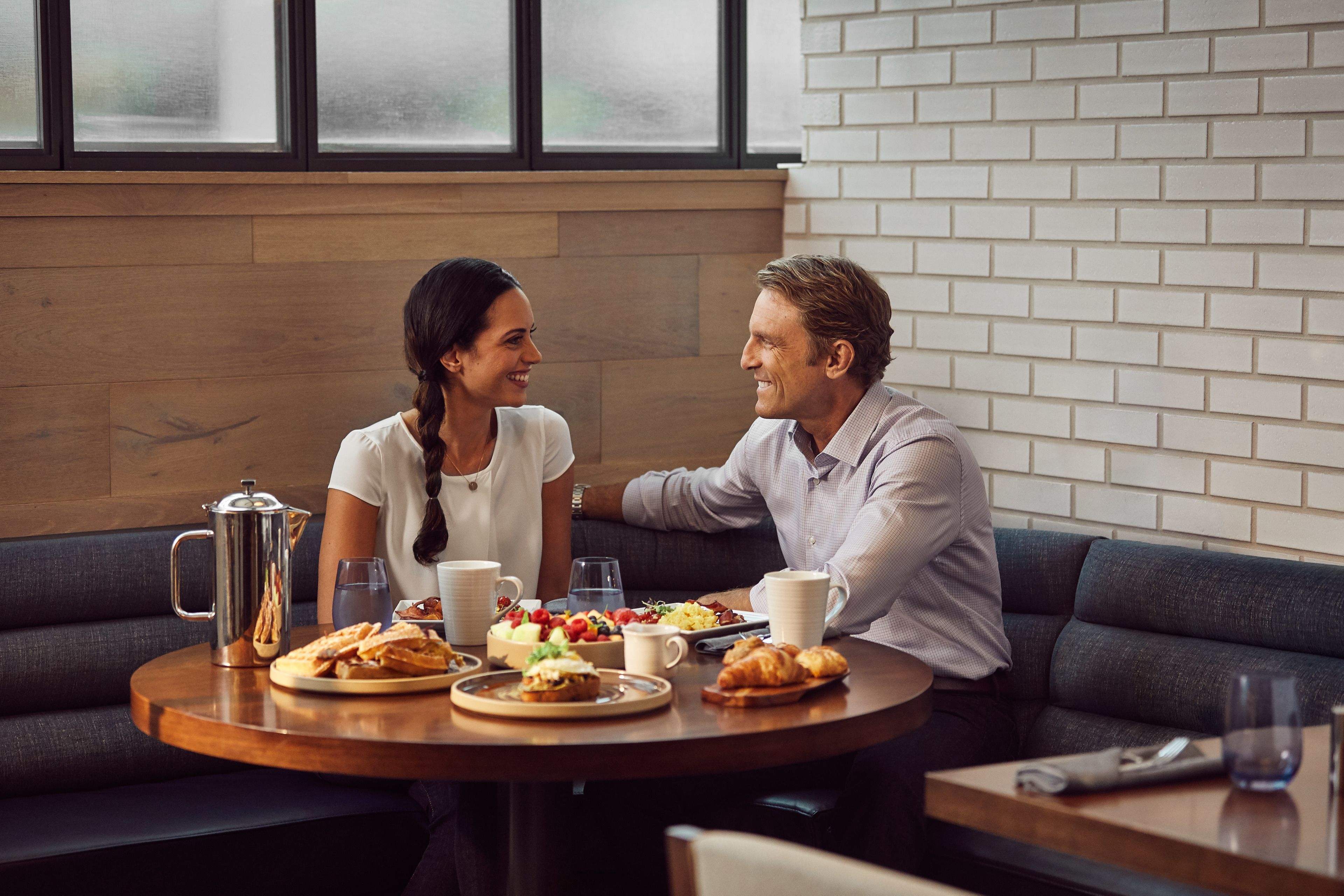 Un couple est assis à une table ronde, devant un petit-déjeuner, une cafetière, et bénéficie d'une vue sur un mur de briques et une fenêtre.