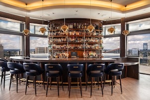 A bar with multiple shelves of bottles, a television, and a city view through large windows.