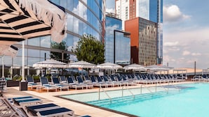 Un área de piscina en la azotea con sillones de descanso y sombrillas, rodeada de edificios modernos.