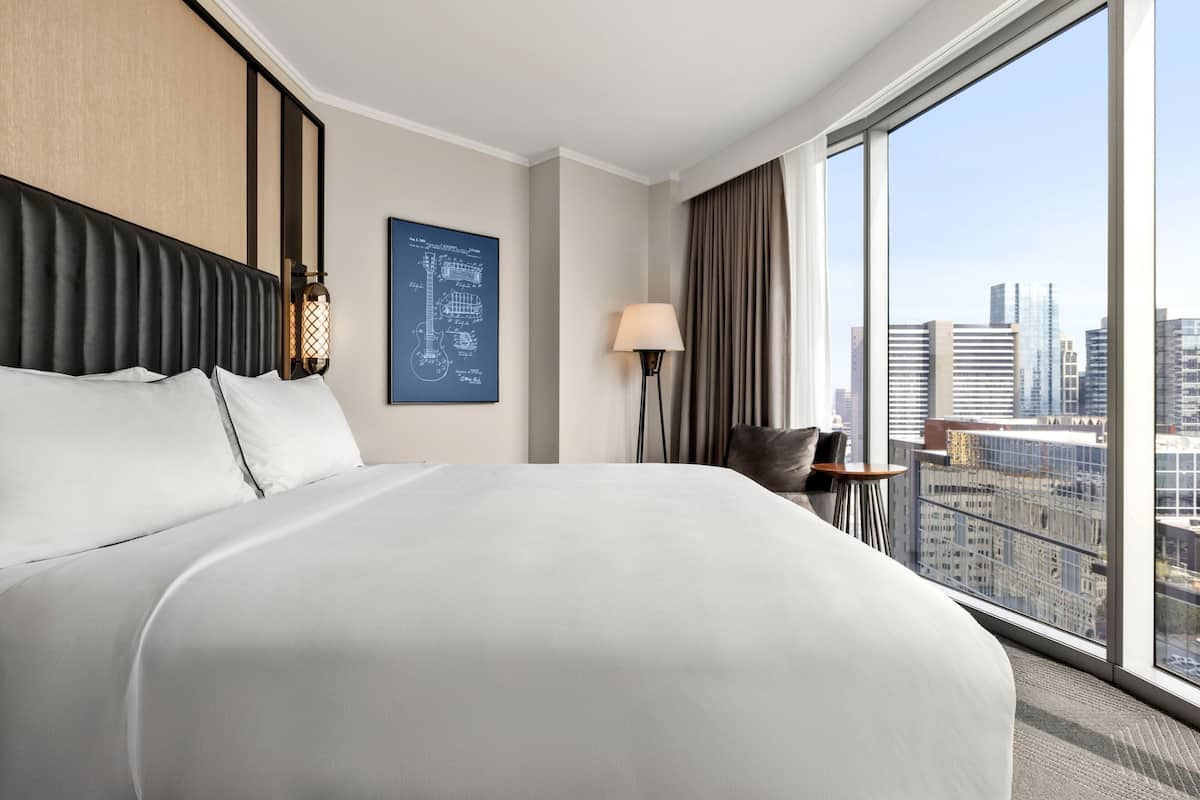 Une chambre d’hôtel moderne avec un grand lit, une vue sur la ville, une lampe et un plan architectural encadré accroché au mur.