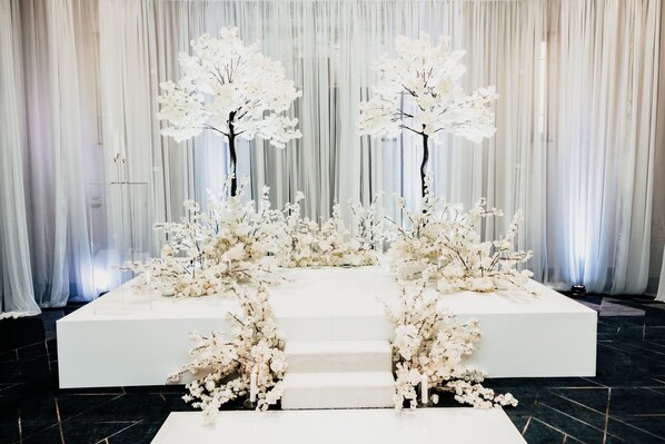 A white reception area with a staircase leading up to a platform, surrounded by white floral arrangements.