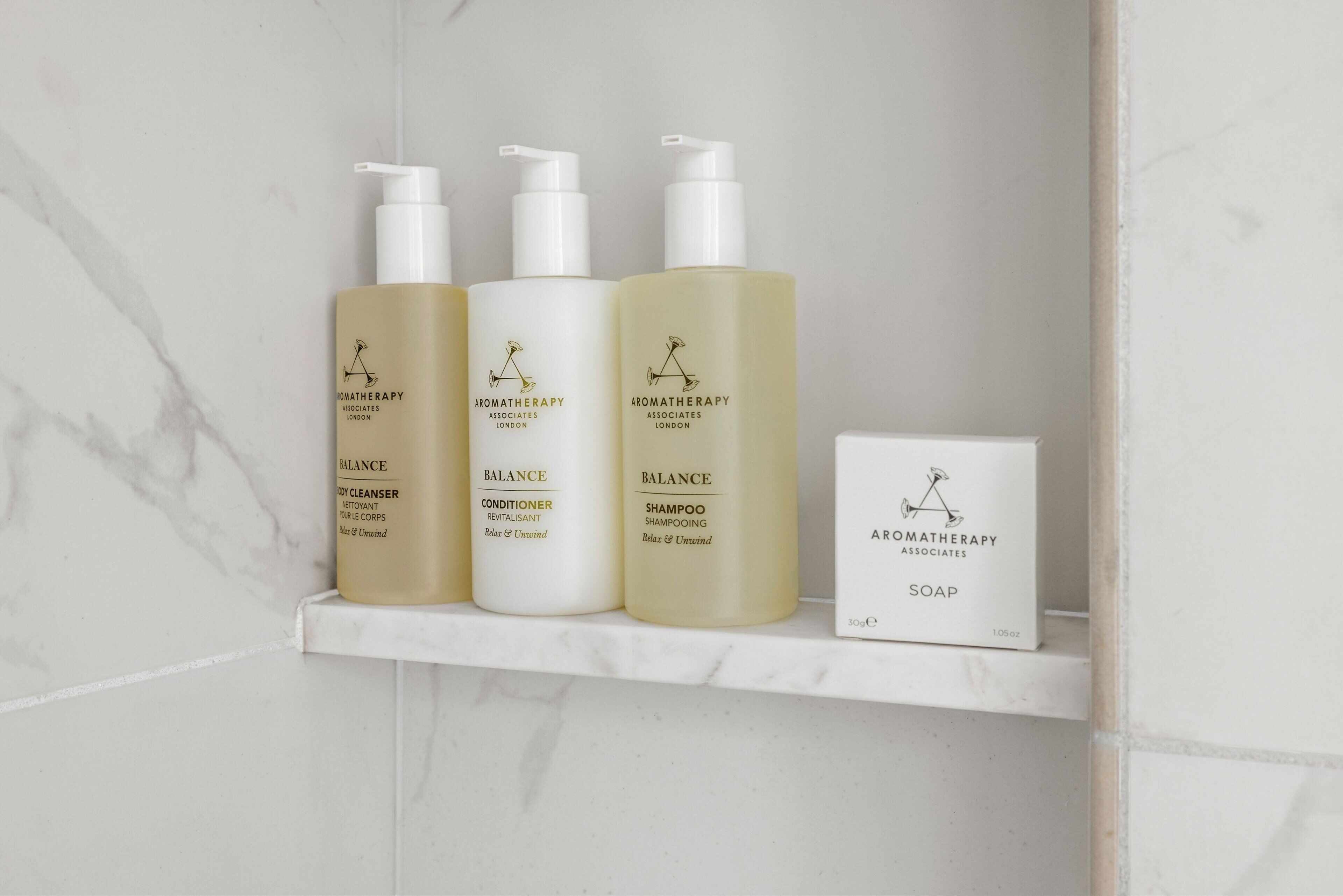 Kylpyhuoneen hyllyllä on kolme Aromatherapy Associates London -tuotemerkkiä olevaa pulloa: vartalopesuaine, hoitoaine ja shampoo, sekä palasaippua.
