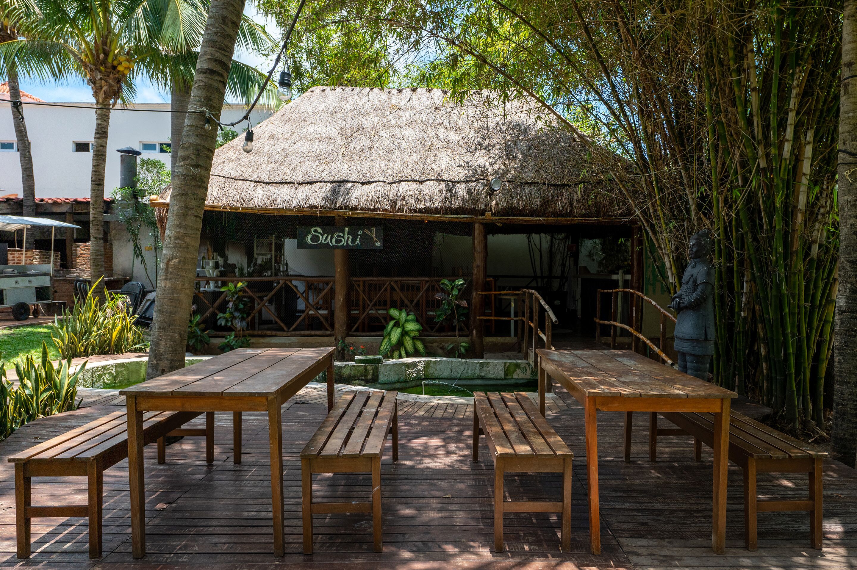 Un área de comedor al aire libre con mesas y bancos de madera, una estructura de techo de paja con la leyenda "Sushi" y un fondo de bambú.