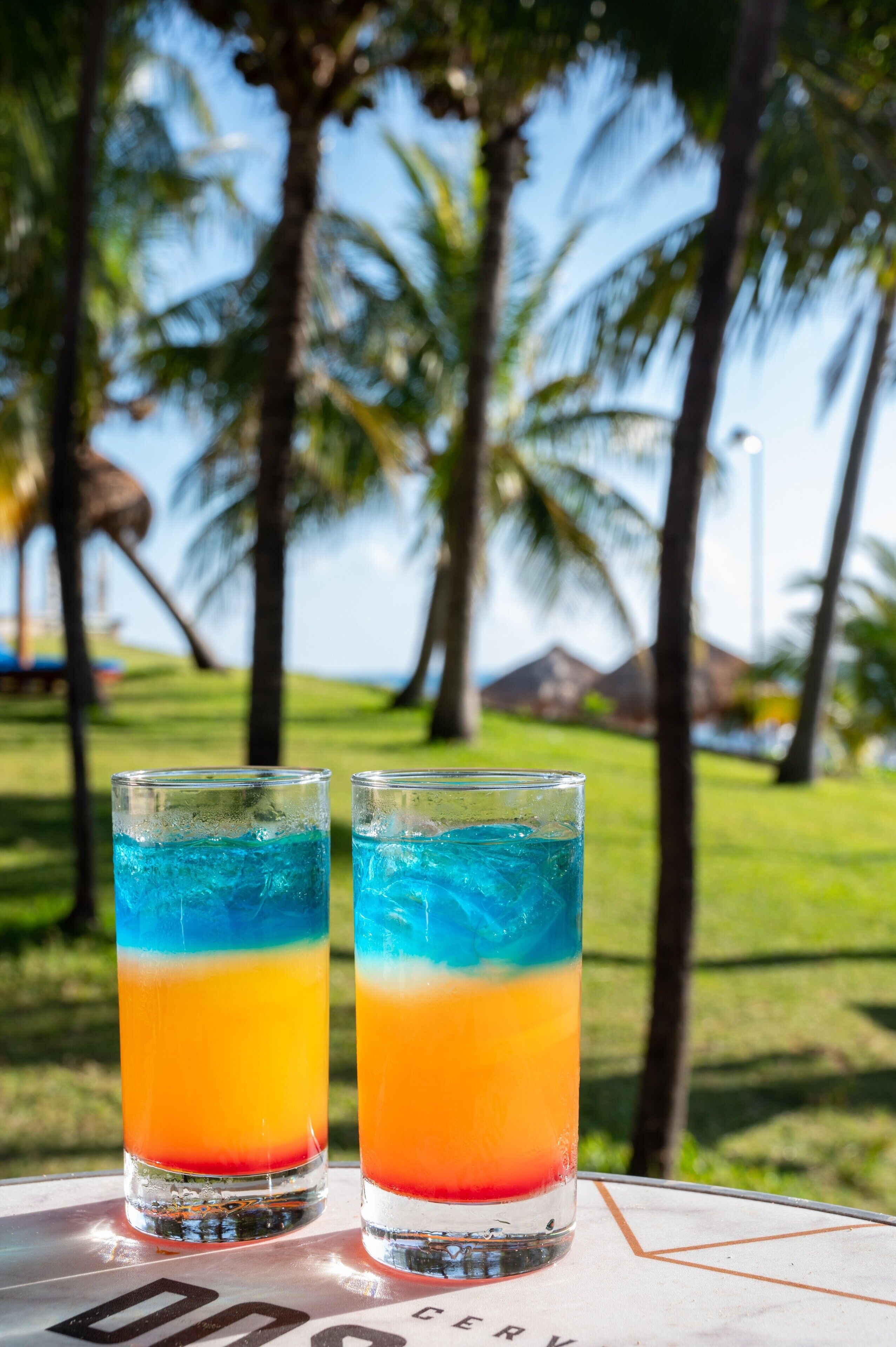 Two glasses of layered drinks on a table with a view of palm trees and a grassy area in the background.