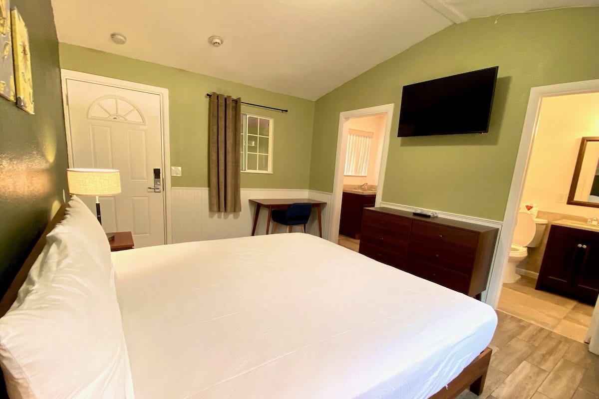 Habitación de hotel con una cama grande, un escritorio, una silla, un televisor y un baño visible a través de una puerta abierta.