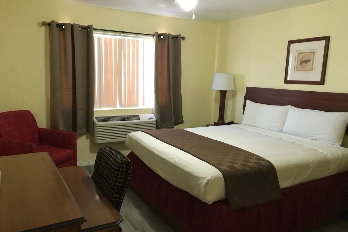 Una habitación de hotel con cama, escritorio, silla, lámpara y un cuadro en la pared.