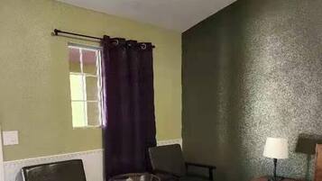 A hotel room with a bed, two chairs, a small table, a window with curtains, and a lamp on a nightstand.
