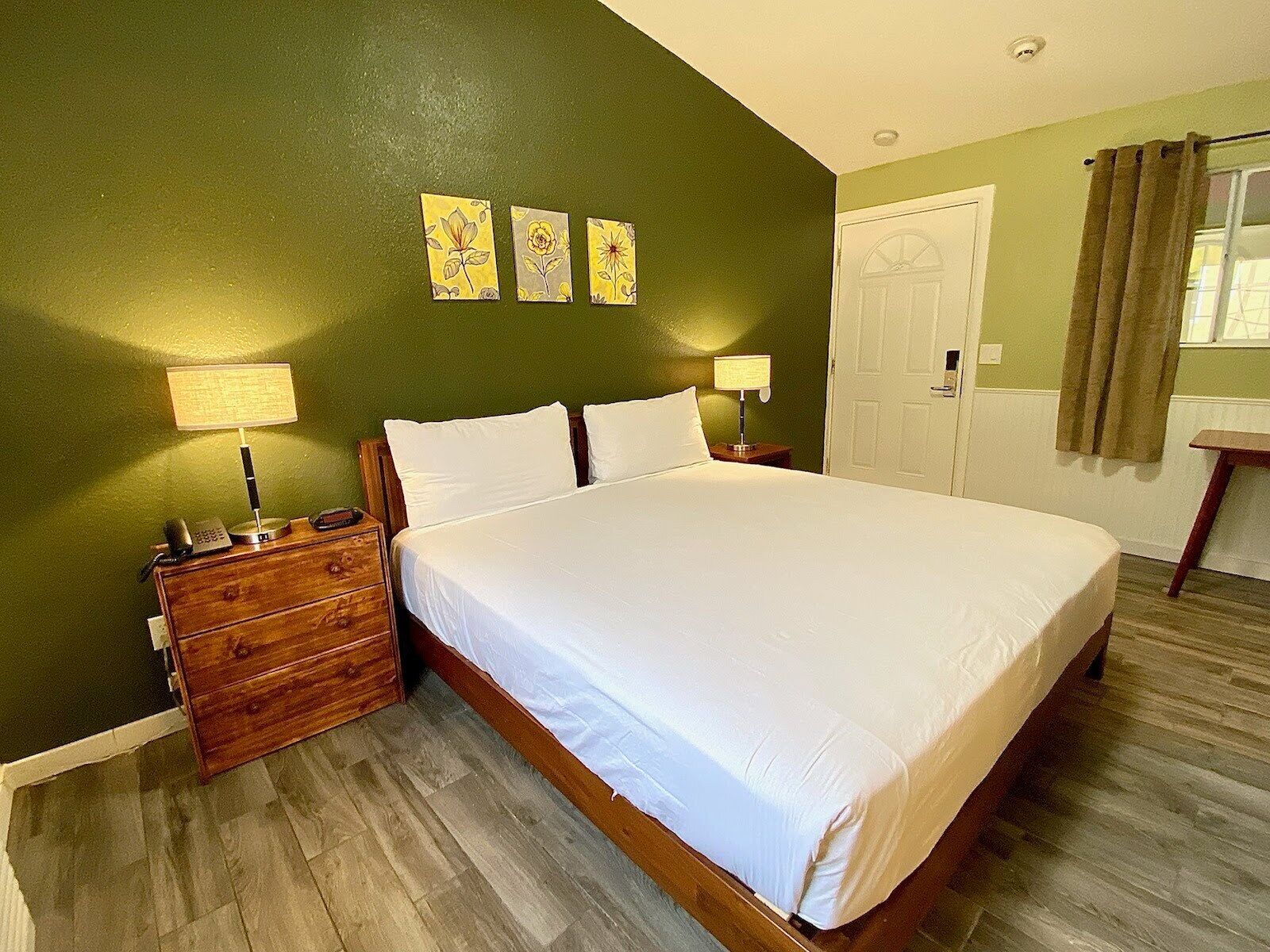 Quarto de hotel com uma cama grande, mesas de cabeceira de madeira, cômoda e uma porta.