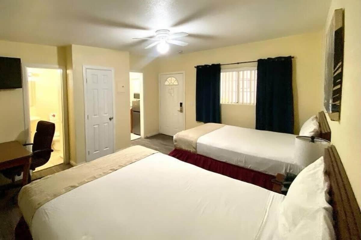 Una habitación de hotel con dos camas individuales, un escritorio con silla, una puerta y un ventanal con cortinas.