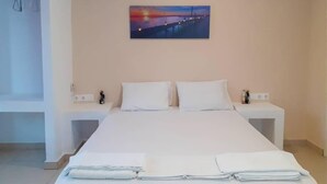 A neatly made bed with white linens and towels, a painting of a sunset over a pier on the wall, and a white nightstand with a lamp. - Sunshine Garden (Hersonissos)