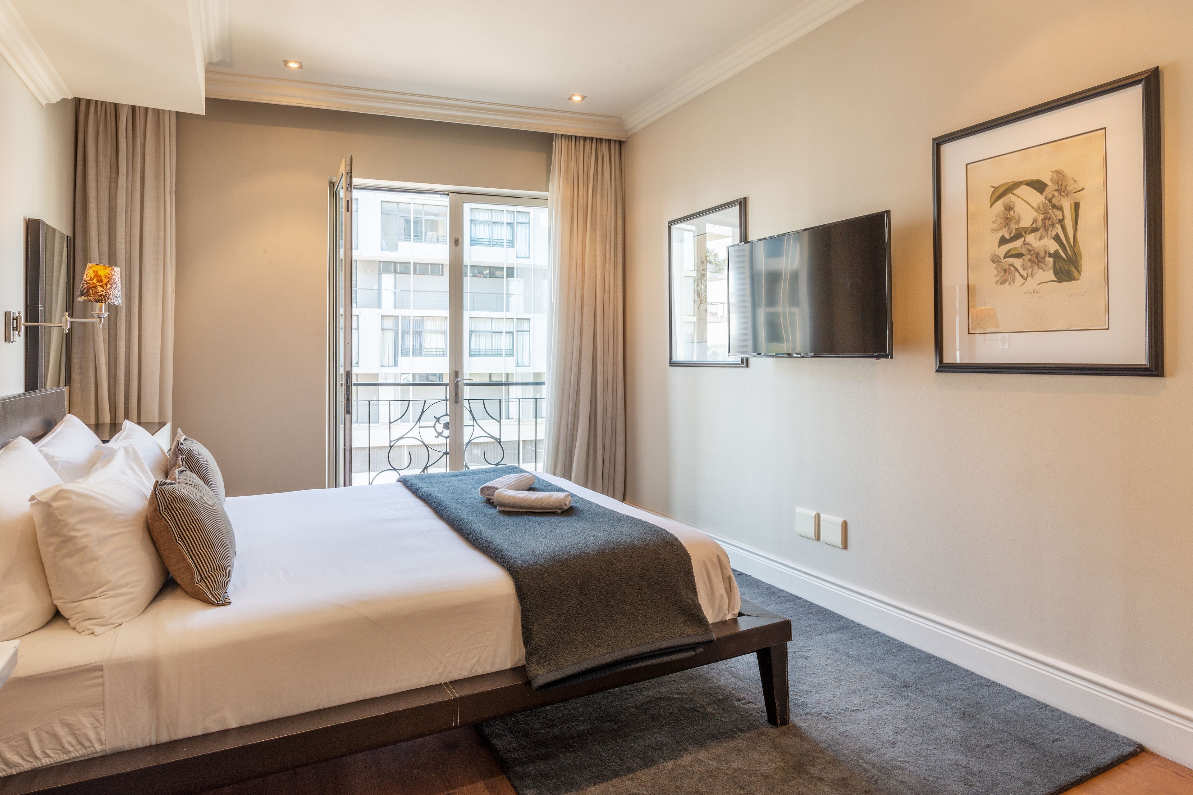 Una habitación de hotel con cama, televisión, un grabado botánico enmarco y balcón con cortinas.