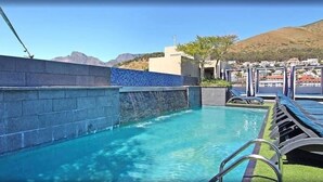 A swimming pool with a stone wall and a blue-tiled section, surrounded by lounge chairs and a view of a hillside town.