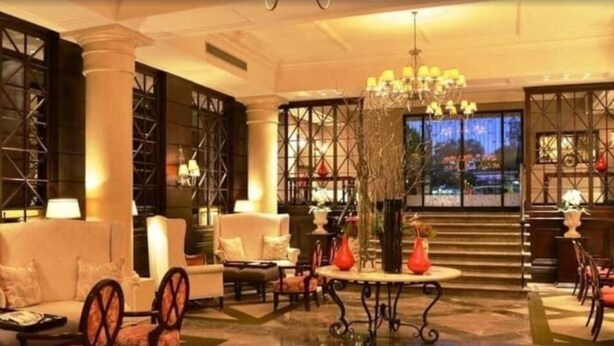 A spacious lobby with a high ceiling, large windows, and a central chandelier. There are multiple seating areas with sofas and armchairs.