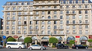 A multi-story hotel with a symmetrical facade, large windows, and balconies. There are several cars parked in front, and a few storefronts with signs visible.