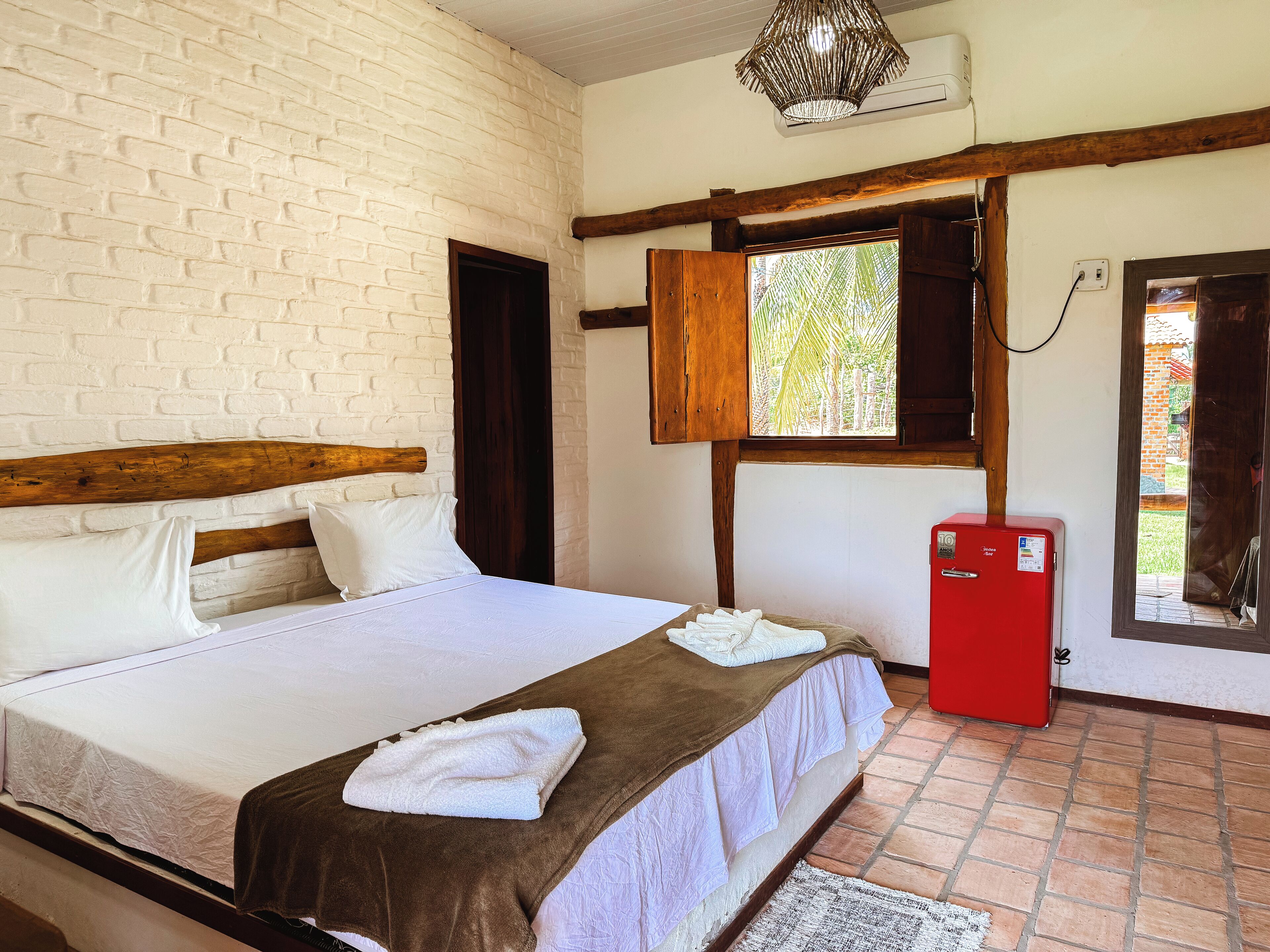 A bedroom with a brick wall, wooden headboard, a bed with white linens, a brown blanket, a red refrigerator, and a window with a view of greenery.