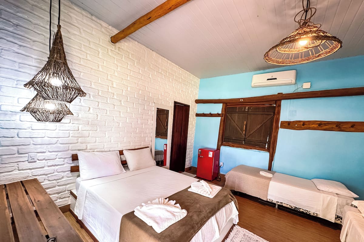 A bedroom with a bed, a bench, a red cooler, and a hanging light fixture.