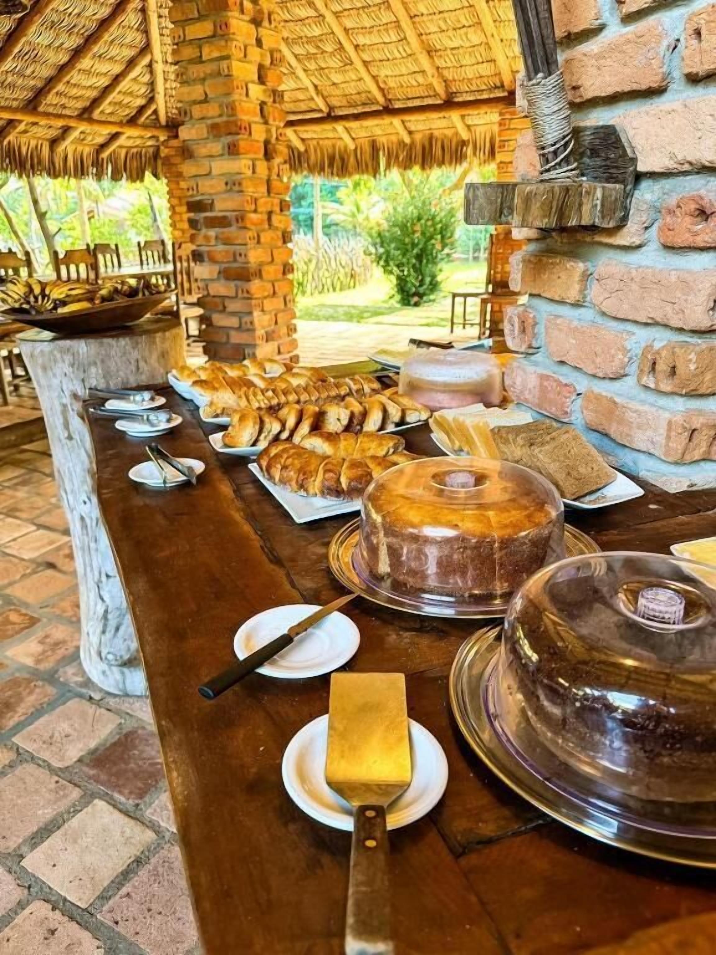 A buffet table with various food items and a cake, set in an outdoor dining area with brick walls and a thatched roof.