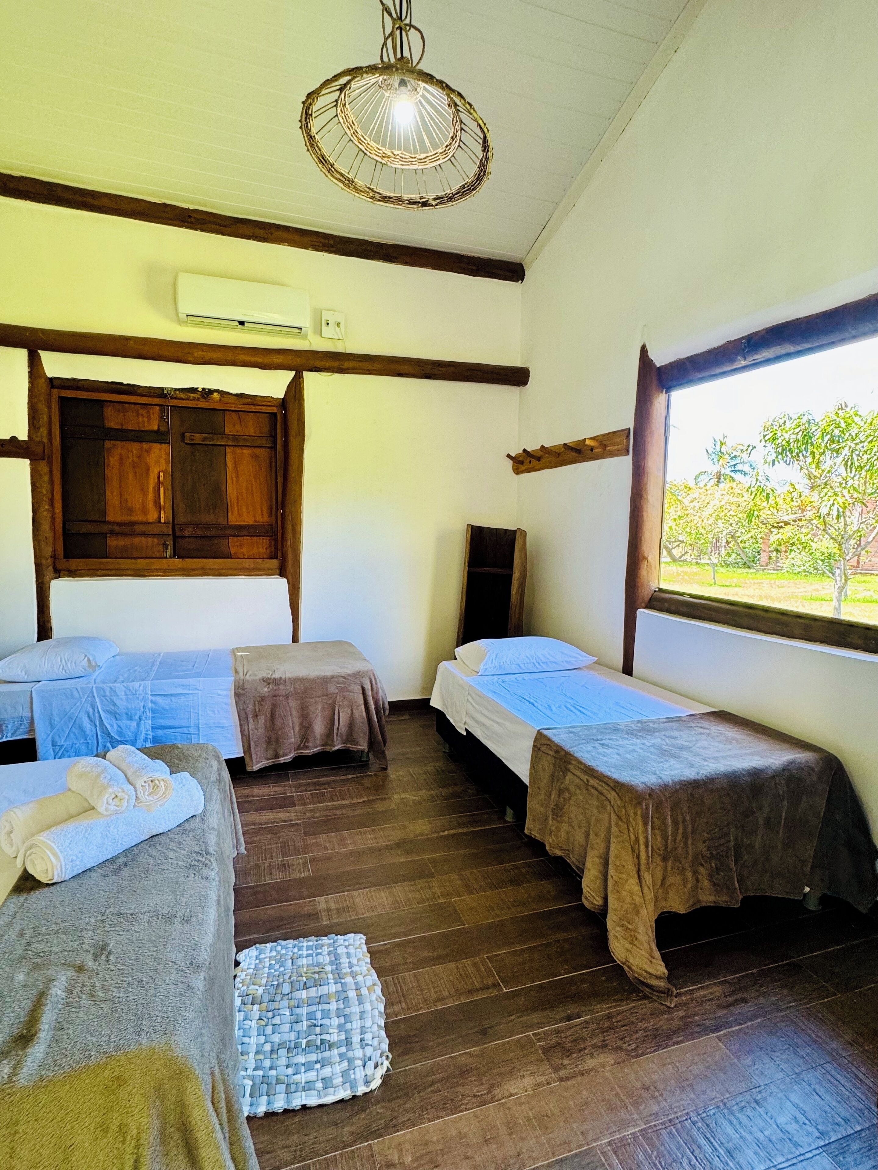 A room with three beds, wooden flooring, a woven ceiling light, and a window with a view of greenery.