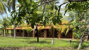 A traditional brick building with a thatched roof, surrounded by lush greenery and a calm water body reflecting the structure. - Pousada Rancho do Buna (Barreirinhas)