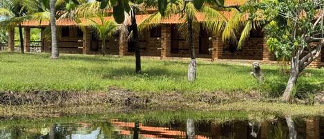 A traditional brick building with a thatched roof, surrounded by lush greenery and a calm water body reflecting the structure.