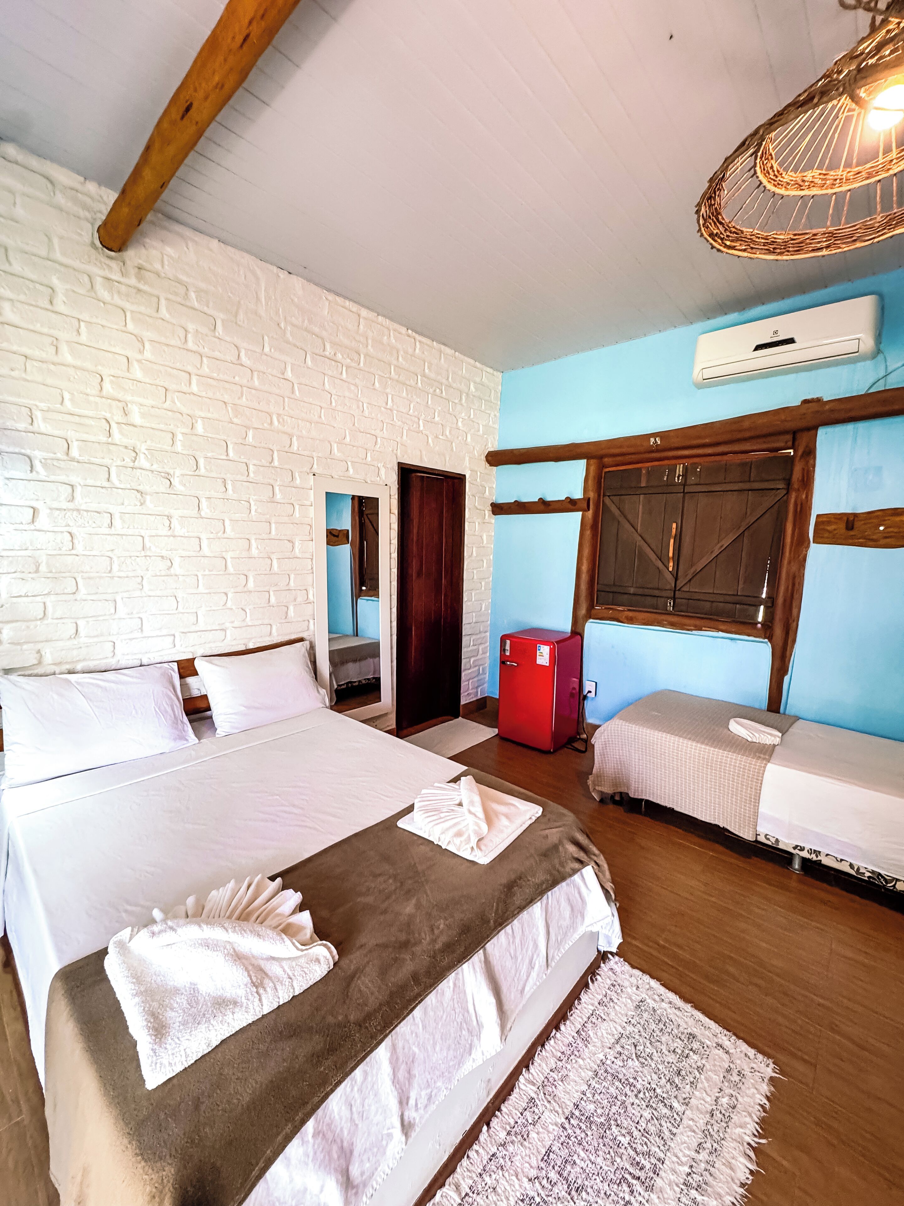 Habitación de hotel con dos camas, un refrigerador rojo, una ventana con contraventanas de madera y una lámpara colgante.