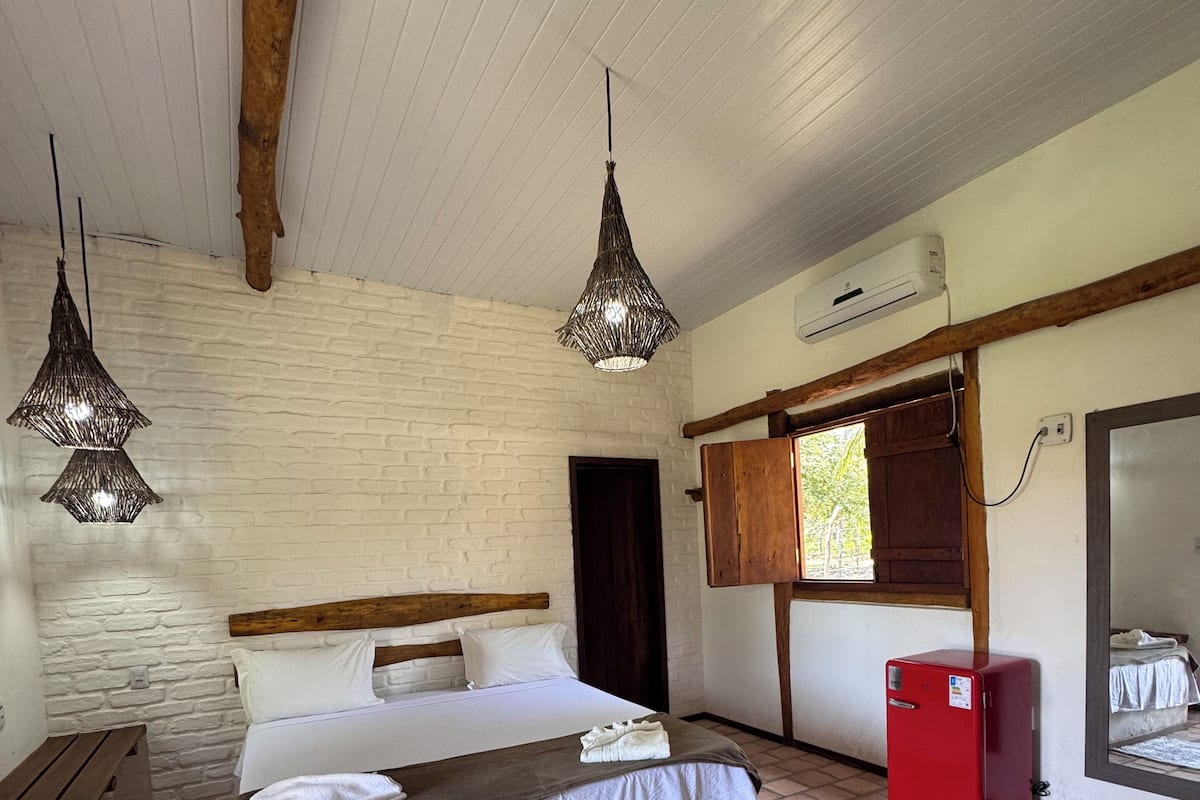 A bedroom with a bed, two hanging light fixtures, a small red refrigerator, a wooden bench, and a brick floor.