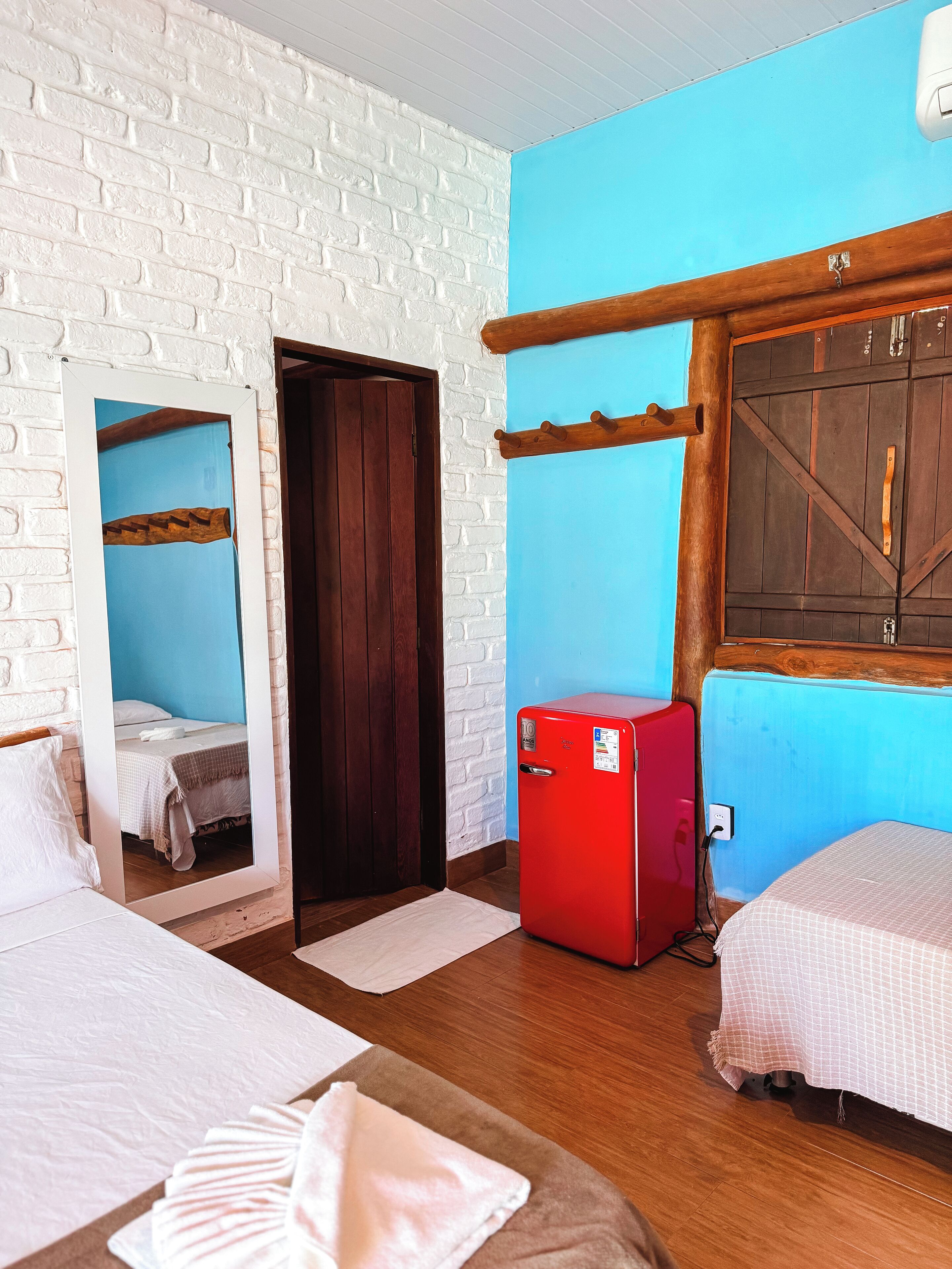 A room with a white brick wall, a wooden door, a red refrigerator, and a bed with a white bedspread.