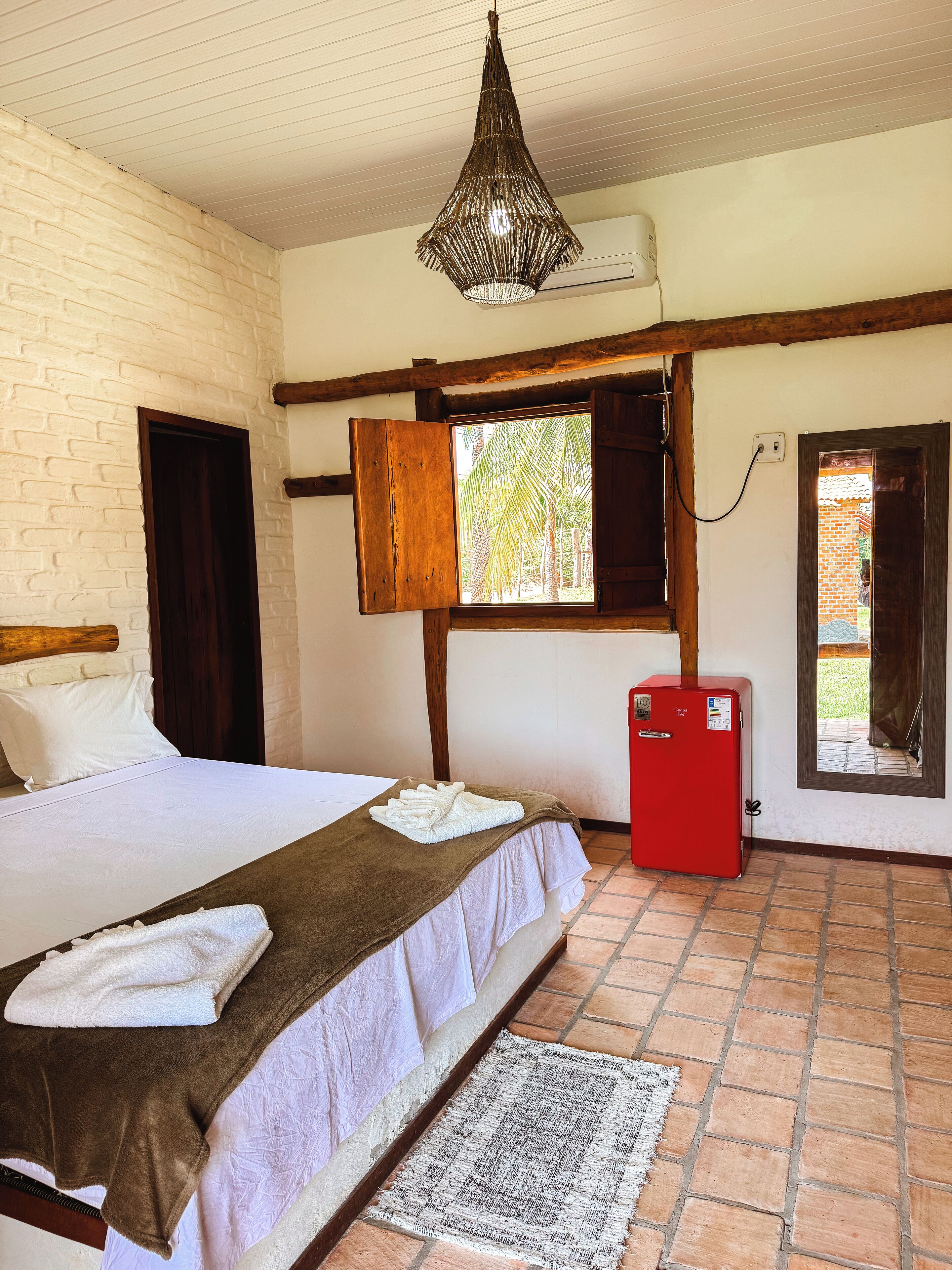 A hotel room with a bed, a red refrigerator, a hanging light fixture, and a window with a view of palm trees.