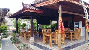 An outdoor seating area with wooden tables and chairs, a thatched roof, and tropical plants. - The Akah Cottage (Lembongan Island)