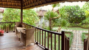A wooden deck with wicker furniture and a thatched roof, overlooking a garden with various plants and a decorative pathway. - The Akah Cottage (Lembongan Island)