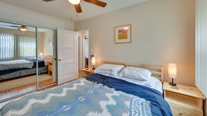 A bedroom with a bed, a ceiling fan, a bedside table with a lamp, a nightstand, a mirror, and a door leading to another room. - Pacific Beach Apartment: Walk to Beach & Pier! (San Diego)