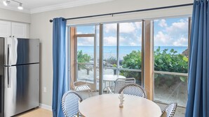 Habitación con vista al océano, una mesa de comedor con sillas, un refrigerador y una ventana con cortinas azules.