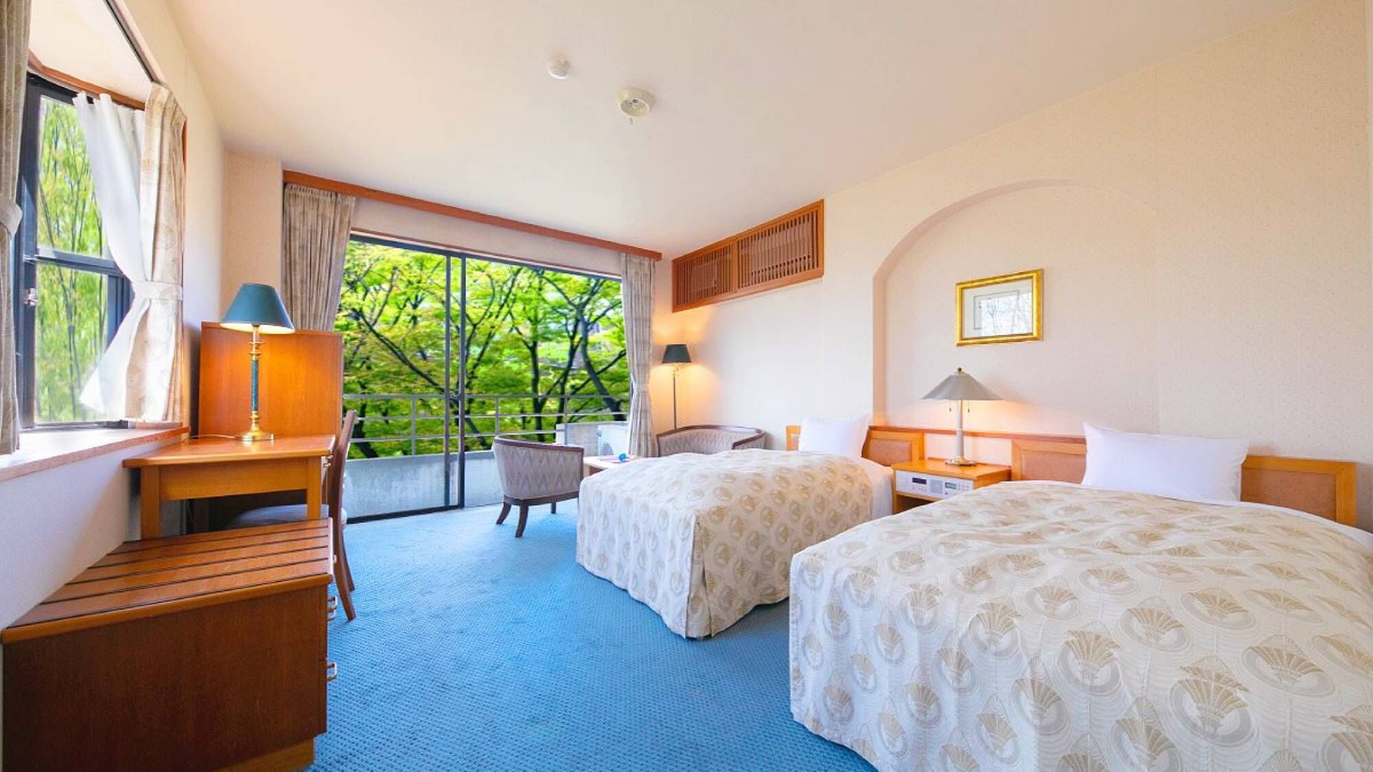 A hotel room with two beds, a desk, a chair, a window with curtains, and a view of trees.
