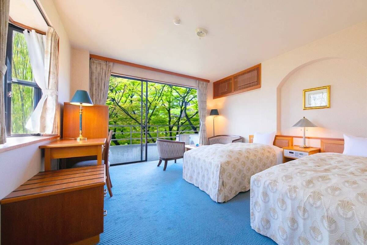 A hotel room with two beds, a desk, a chair, a window with curtains, and a view of trees.