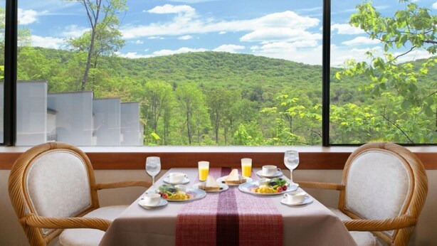 Una mesa de comedor con platos, vasos y mantel rojo, frente a una vista de una ladera boscosa.