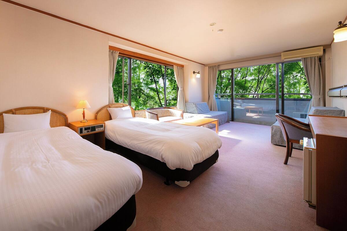 A hotel room with two beds, a desk, a chair, and a balcony with a view of trees.