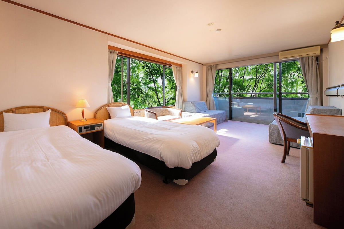 A hotel room with two beds, a desk, a chair, and a balcony with a view of trees.