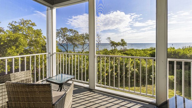 A covered balcony with wicker furniture and a glass table, overlooking a body of water and greenery. - Cedars East Bayview Tennis & Pool Unit 822 by RVA (Longboat Key)